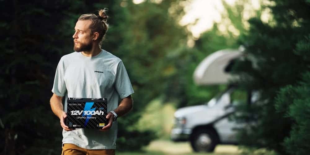 A man carries a 12V battery in a green outdoor setting