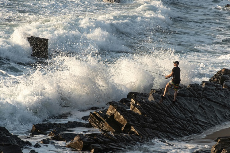 A person fishing on a rocky shoreline with powerful waves